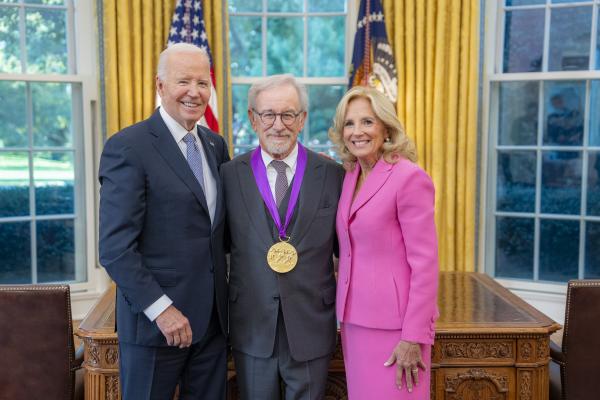 Man with glasses and gray hair and beard in gray suit wearing a medal around his neck surrounded by older man wearing a blue suit and woman with blonde hair wearing a pink suit.