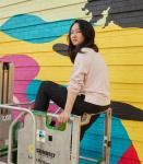 Asian women on scaffolding in front of mural she is painting. 