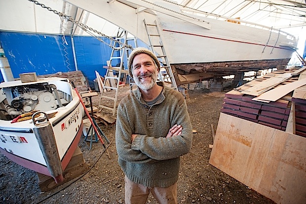 A man poses in front of a sailboat hoisted on props in a workshop.